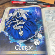Load image into Gallery viewer, A blue and silver dice bag is closed on a gaming table with a dice tray and D&D books in the background.