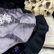 Load image into Gallery viewer, A silver and black dice bag is open flat on D&D gaming books with skulls in the background.