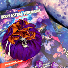 Load image into Gallery viewer, A purple and orange dice bag with a metal planet charm sits closed on a stack of D&D books.