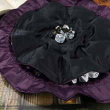Load image into Gallery viewer, A black and purple dice bags is opened flat on a gaming table. There are black, white, and gold dice in the bag.