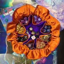 Load image into Gallery viewer, A purple and orange dice bag sits open on a gaming table. Inside, the pockets are filled with colorful dice.