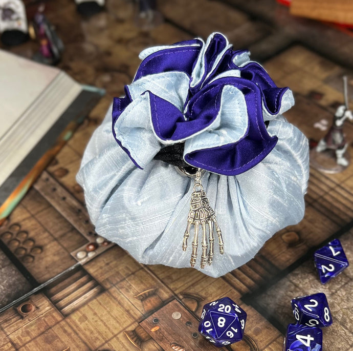 A light blue and purple dice bag with a metal skeletal hand charm sits closed on a gaming table.