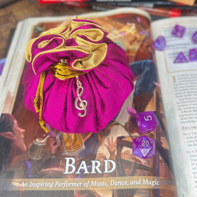 A pink and gold dice bag is closed on a gaming table with a dice tray and D&D books in the background.