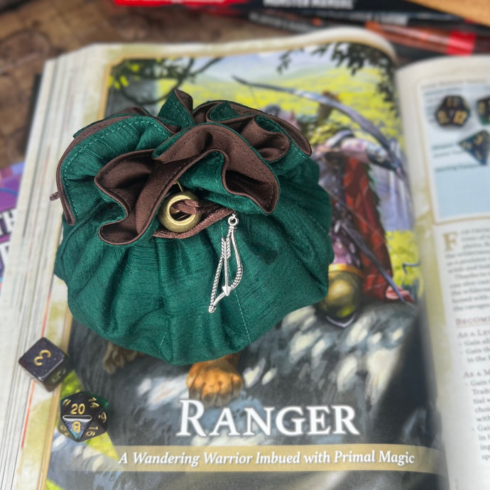 A green and brown dice bag is closed on a gaming table with a dice tray and D&D books in the background.