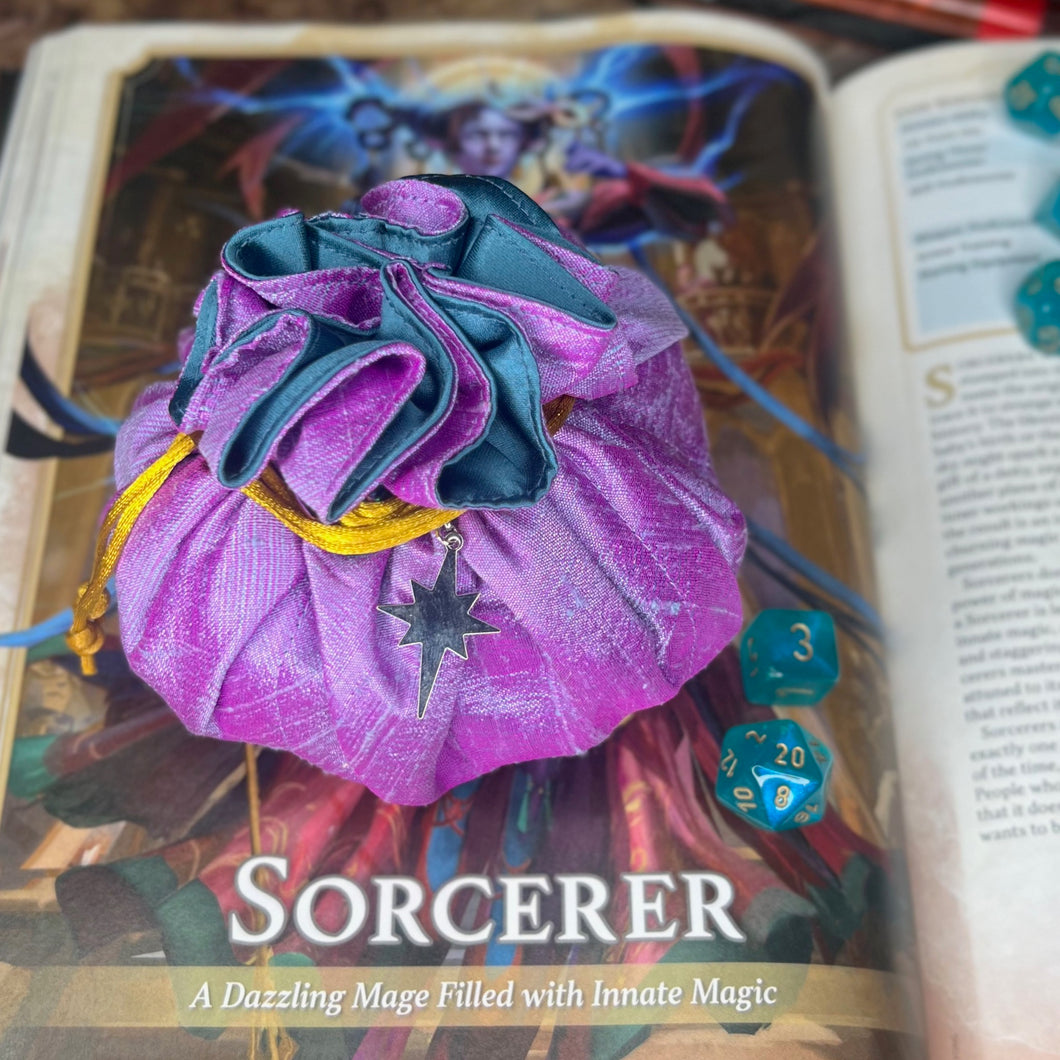 A purple and blue dice bag is closed on a gaming table with a dice tray and D&D books in the background.