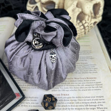 Load image into Gallery viewer, A silver and black dice bag with a metal skull charm sits closed on top of an open D&amp;D book.
