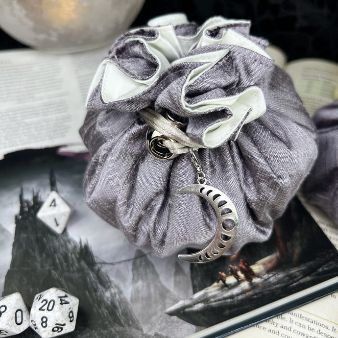 A silver and white dice bag with a silver moon charm sits closed on a gaming table.