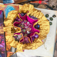 Load image into Gallery viewer, A pink and gold dice bag is open on a gaming table with a dice tray and D&amp;D books in the background.
