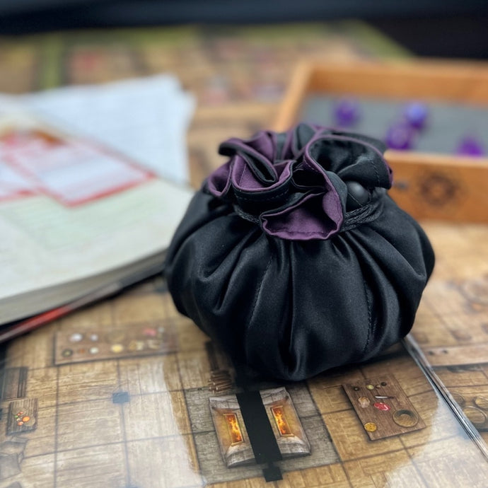 A black and purple dice bag sits closed in front of a hardwood dice tray.