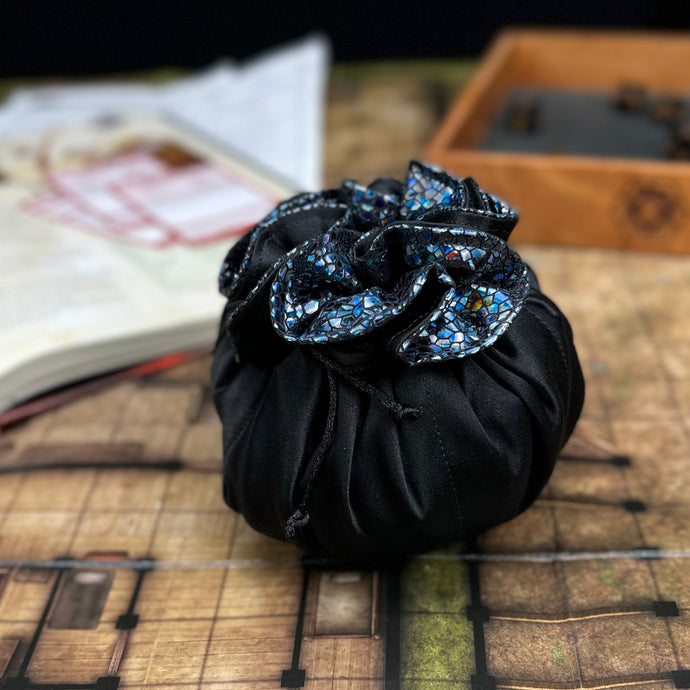 A black and shimmering dice bag sits closed on a gaming table with a dice tray and book in the background.